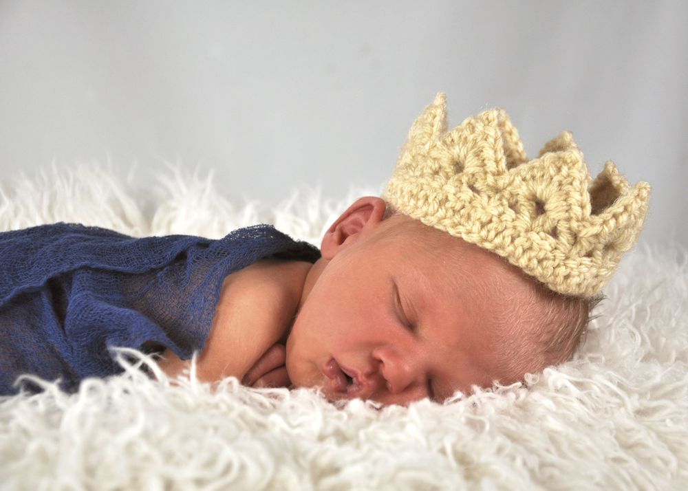 Sleeping baby wearing a crocheted golden crown on white fur blanket.