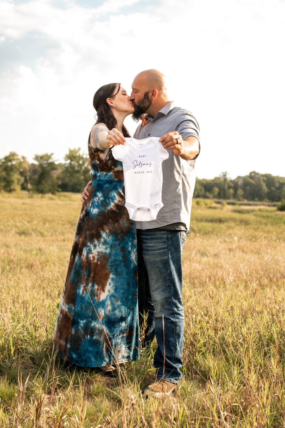Couple embracing in field while holding baby announcement onesie.