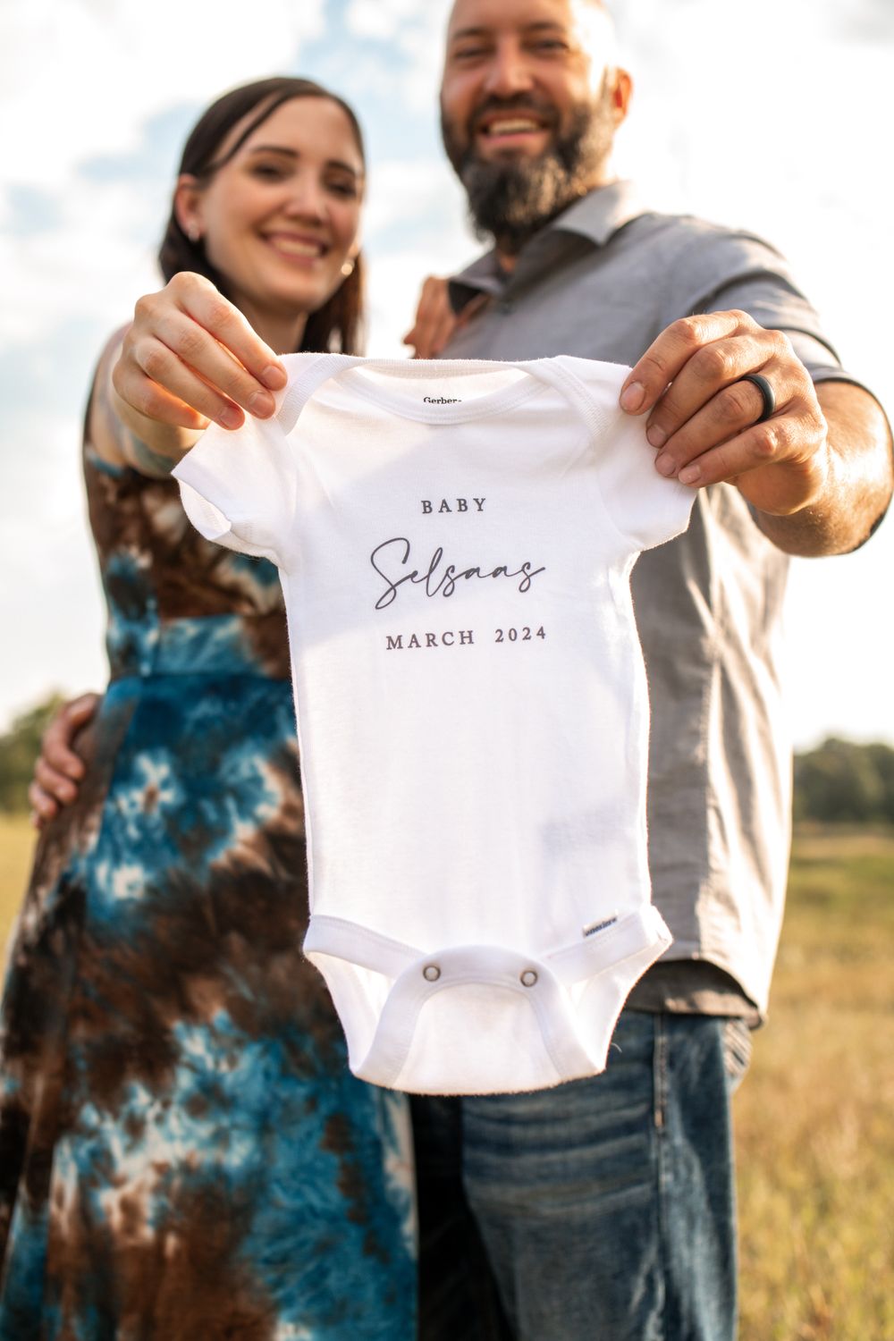 Expectant couple displaying white baby onesie with 'March 2024' text.