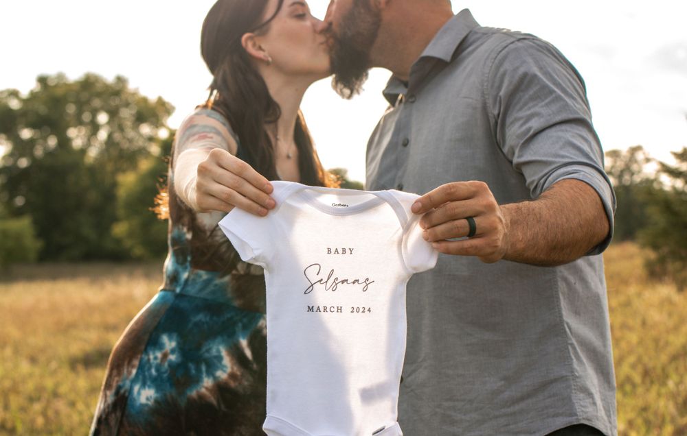 Couple holding a white baby onesie in golden field during sunset.