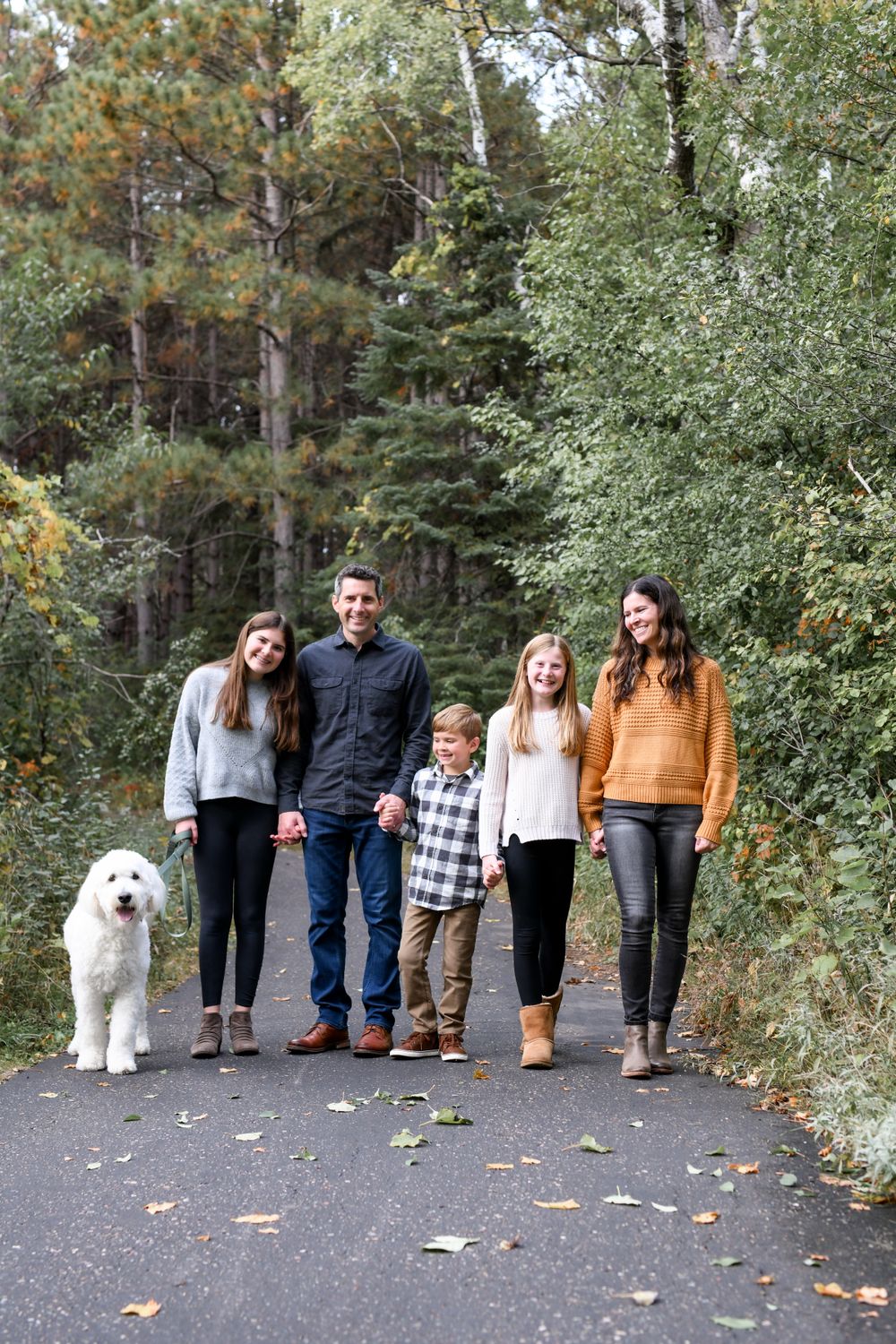 Family photo session on path with white dog against autumn woodland backdrop.