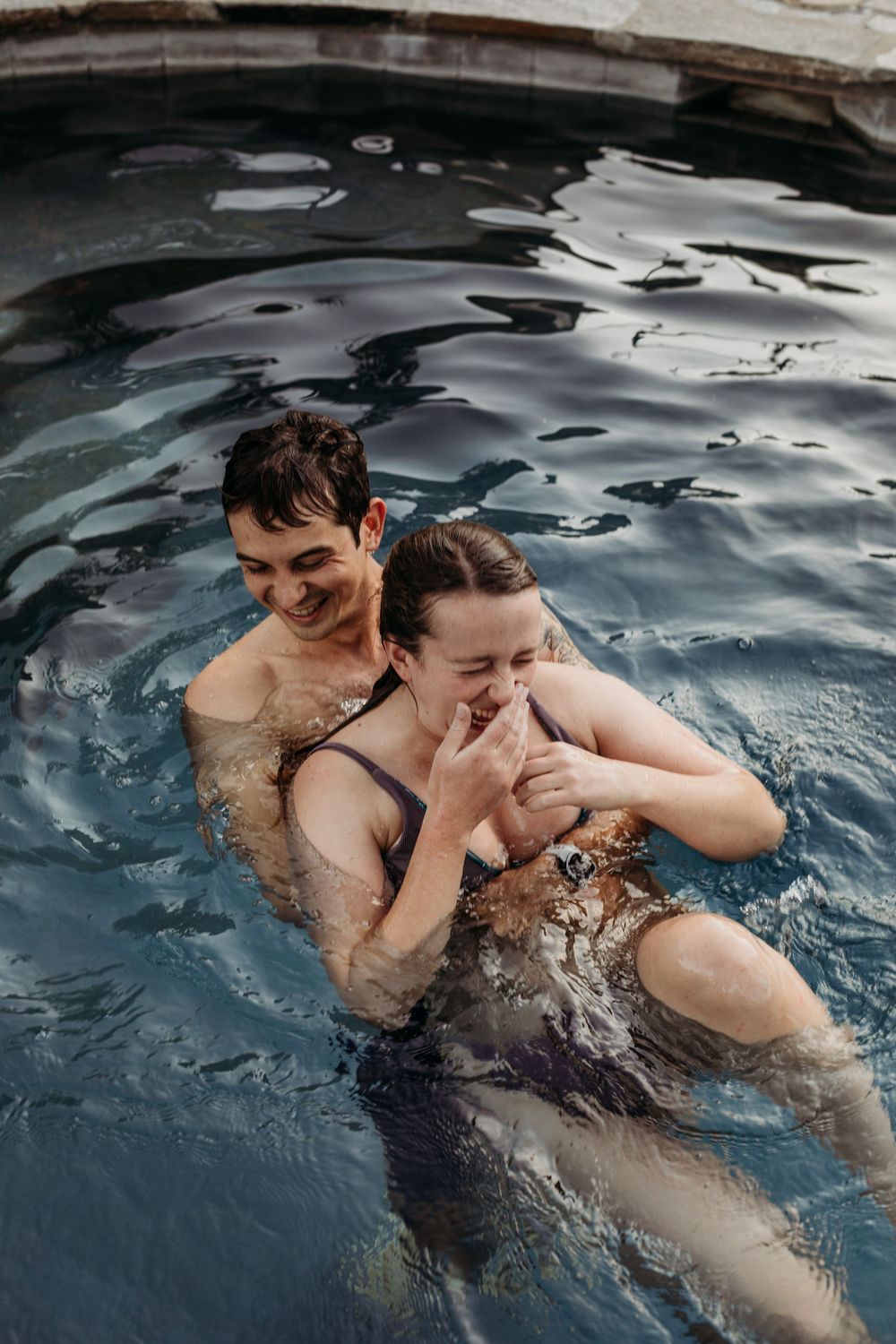 Two people share laughter while swimming in a pool on a cloudy day.