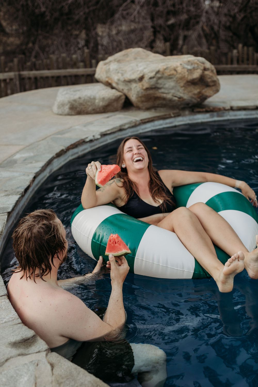 Friends enjoying watermelon while floating in a swimming pool with rock features.