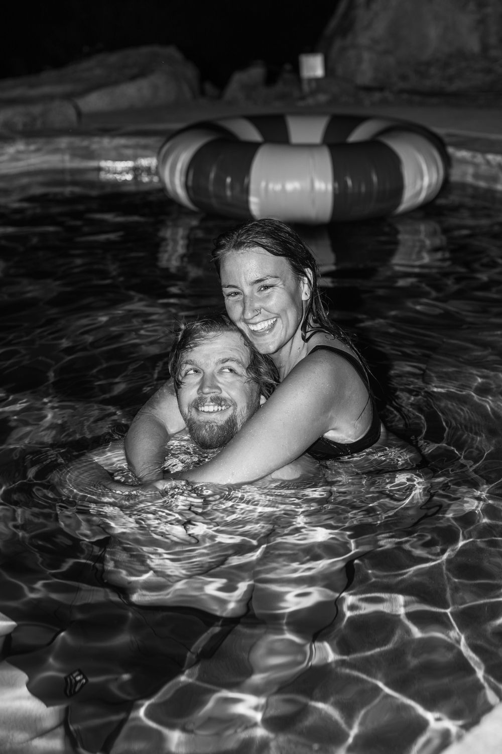 A couple embraces while swimming in a nighttime pool with an inflatable ring float.