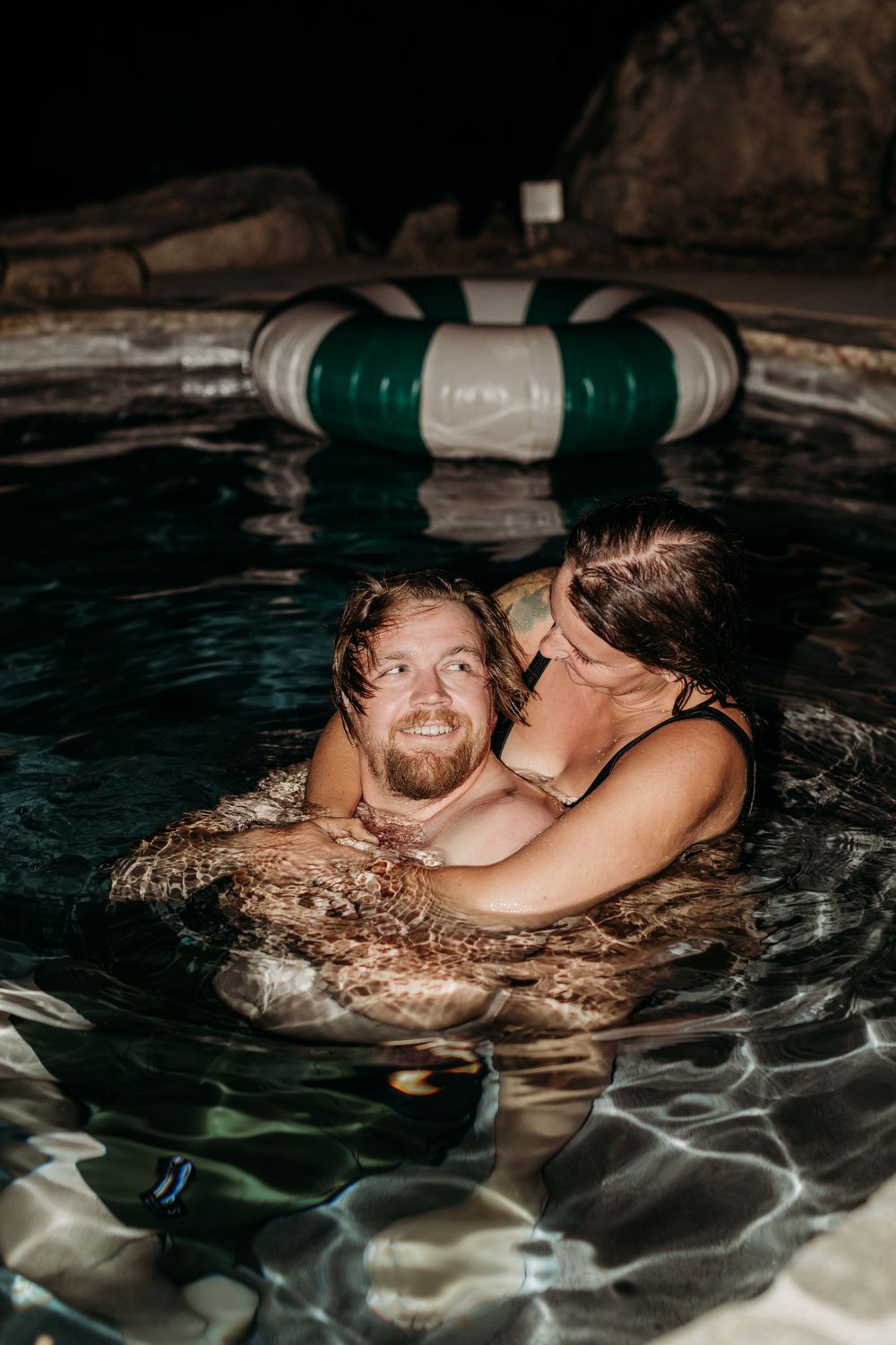 Two people share an intimate moment while swimming in a dark pool with a striped float.