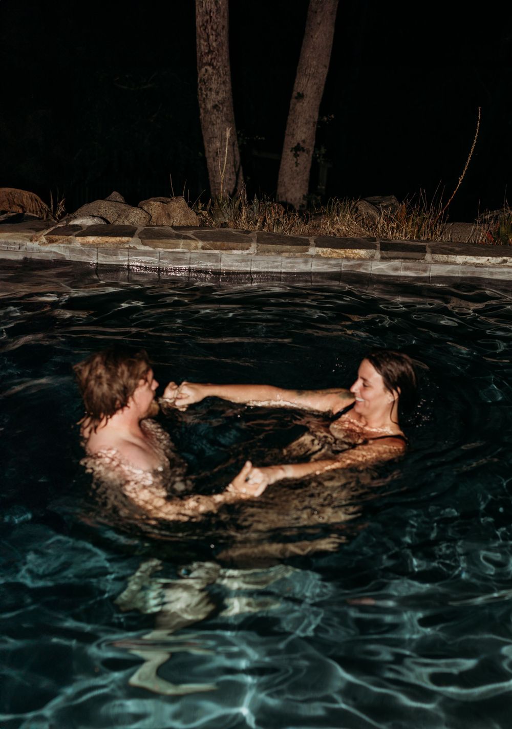 Two swimmers playfully interact in a nighttime pool surrounded by rocks and trees.