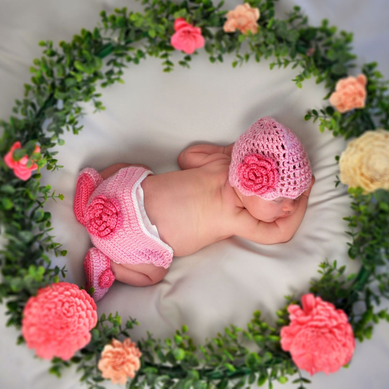 Newborn baby sleeping in pink outfit on lace backdrop with floral decorations.