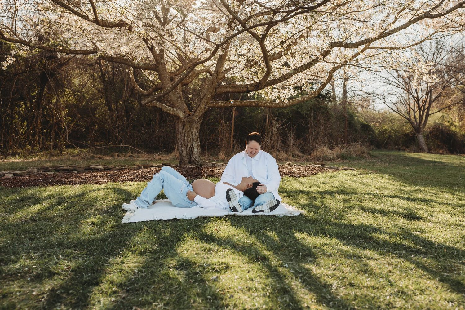 Expecting couple relaxing on blanket under blooming tree in sunny green park.