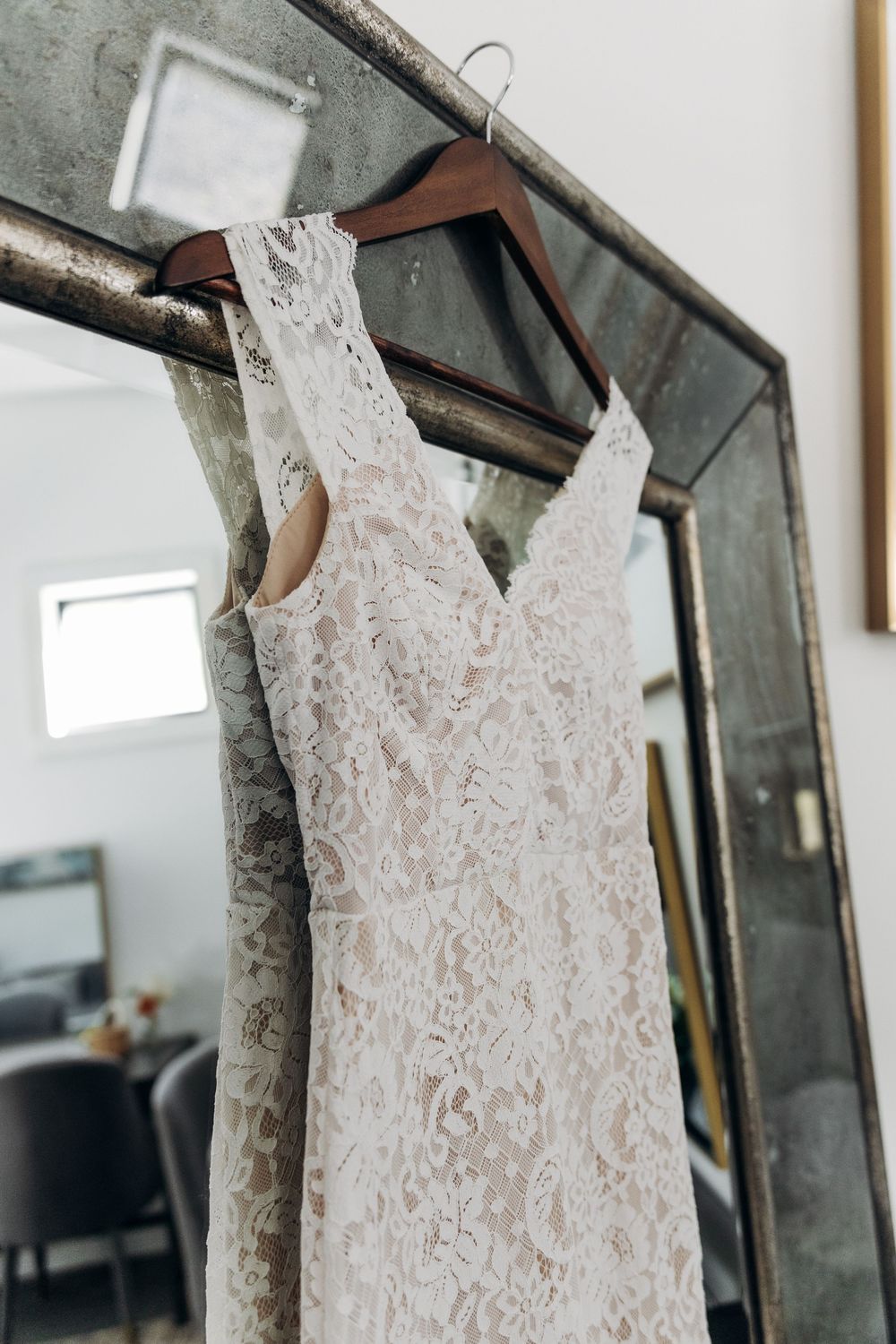 A delicate white lace wedding dress hangs on a dark wooden hanger against a light colored wall.