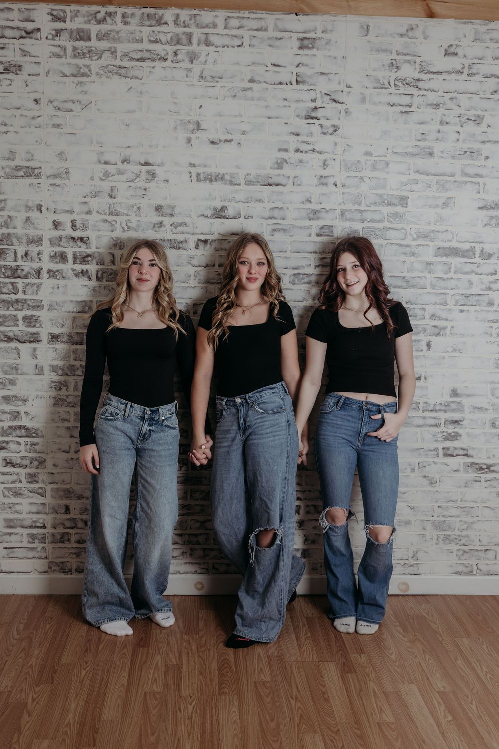 Three people in matching black tops and blue jeans sit casually against a white brick wall.