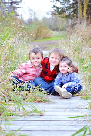 Three children sitting together on wooden boardwalk surrounded by tall grass.