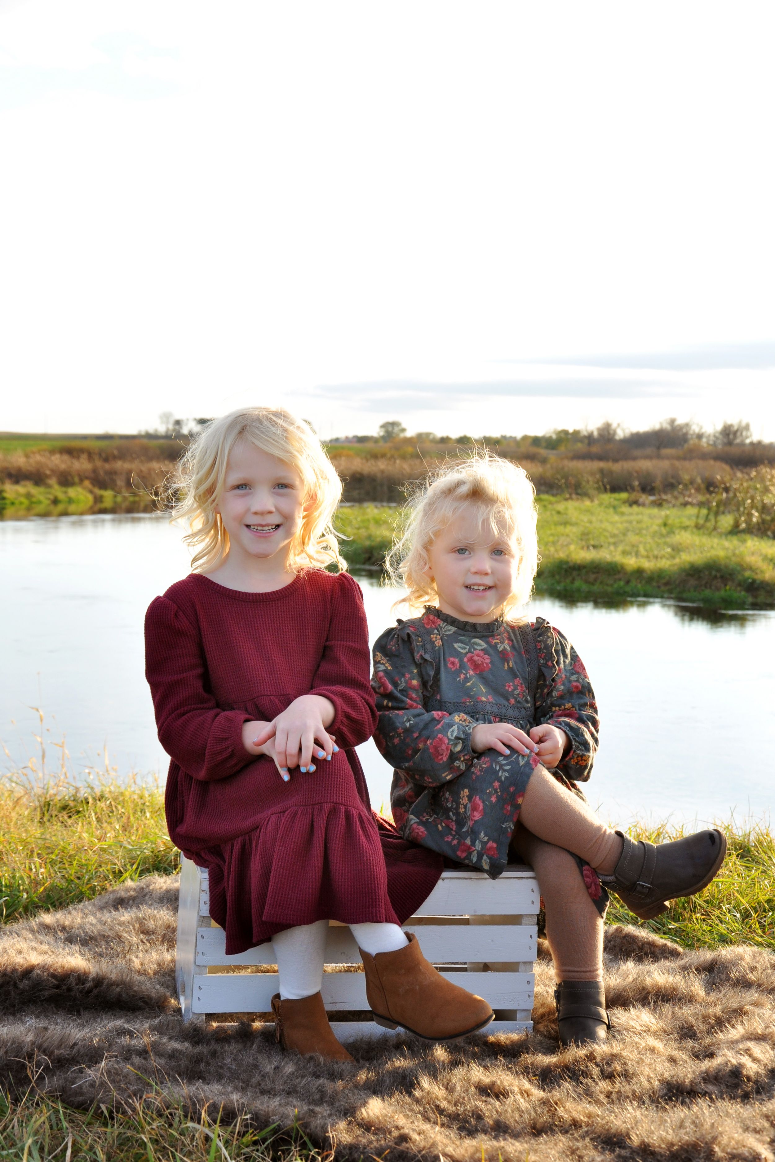 Two children sitting on white wooden crate outdoors near water at sunset.