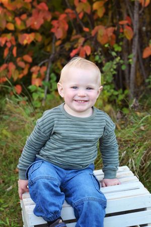 Child in striped sweater and jeans sitting on white wooden crate with fall foliage background.