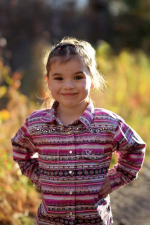 Child in pink patterned shirt standing outdoors with sunlit autumn background.