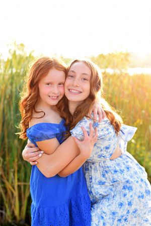 Two children in blue and floral dresses hug while posing in golden sunset light.