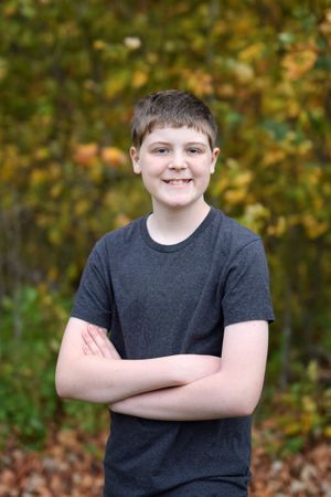 Child in dark t-shirt poses with arms crossed against autumn foliage background.
