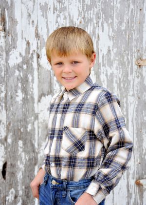 Young person in plaid shirt and jeans poses playfully against weathered white wood background.