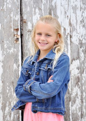 Child in denim jacket strikes different poses against a distressed white wooden wall.