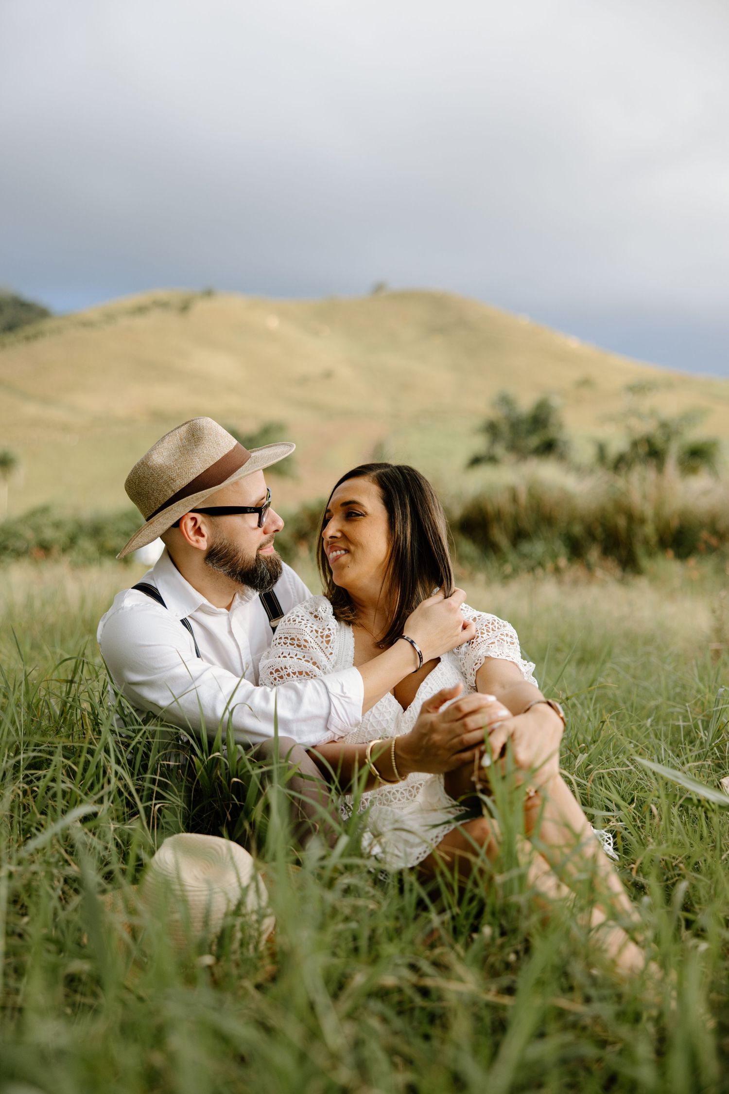 Romantic couple embracing while sitting in tall grass with rolling hills in background.