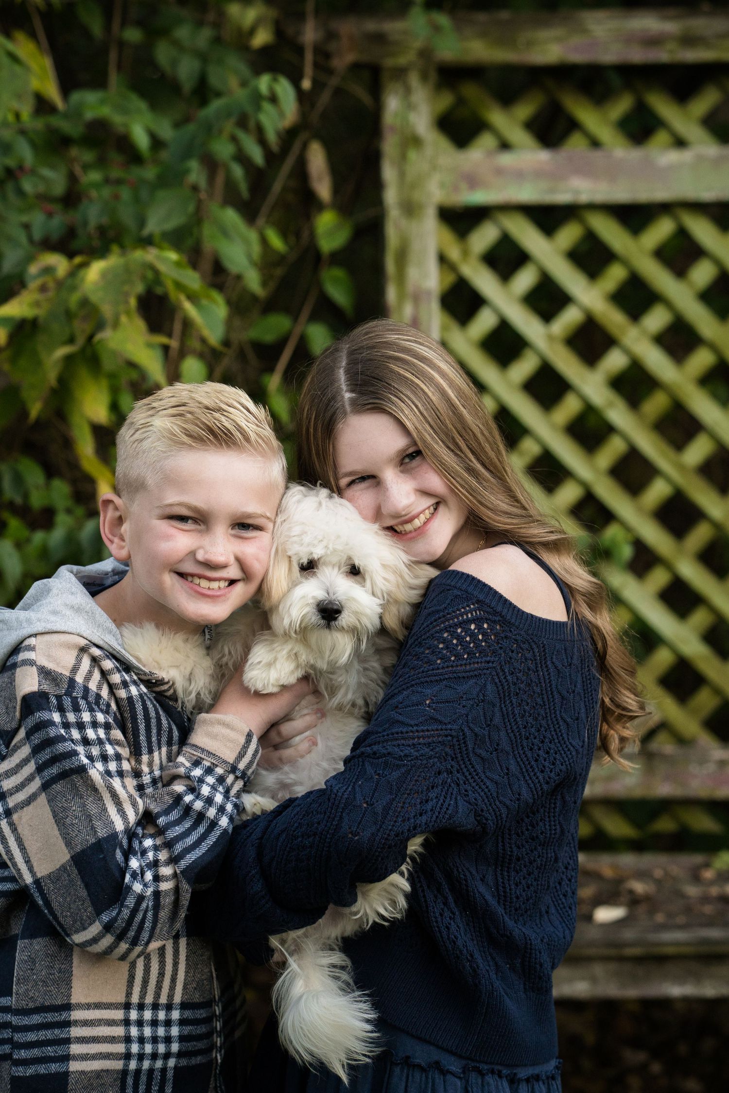 Two smiling kids holding a fluffy white dog outdoors near a wooden lattice fence surrounded by green foliage.