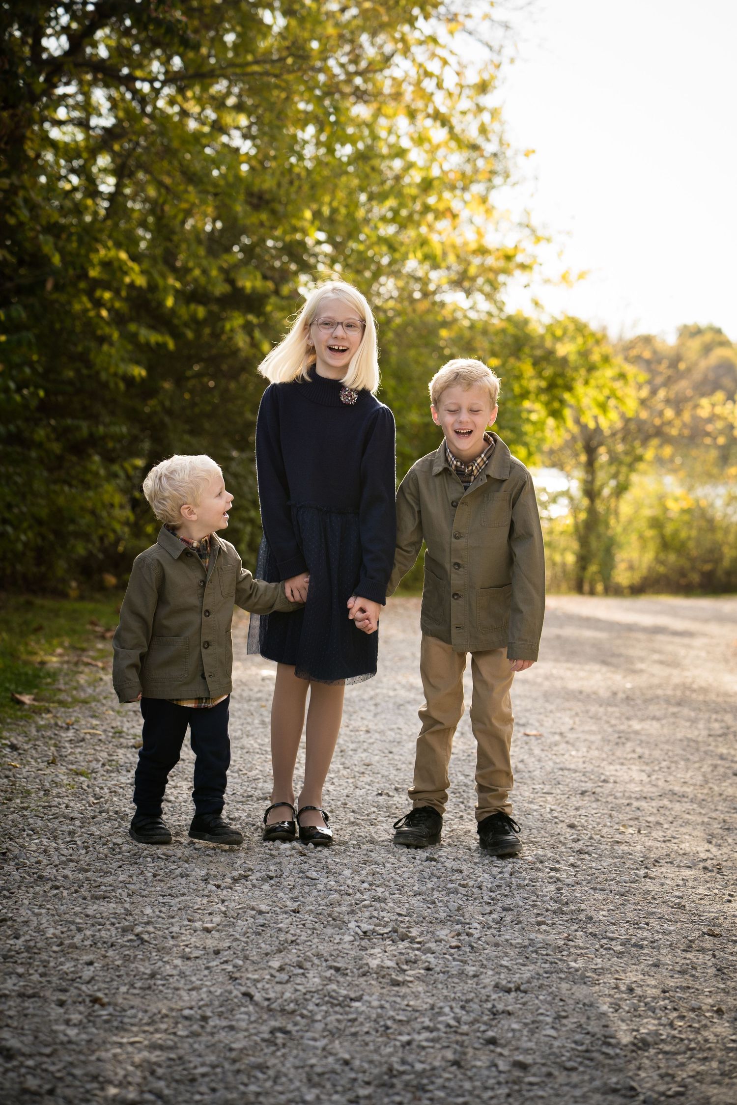 Three happy children holding hands on a gravel path surrounded by autumn trees in golden light.