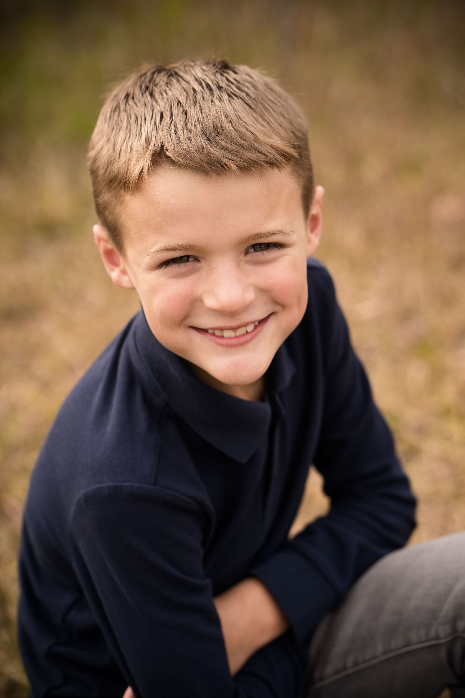 Smiling young boy in navy blue shirt sitting outdoors in a field, portrait photo.