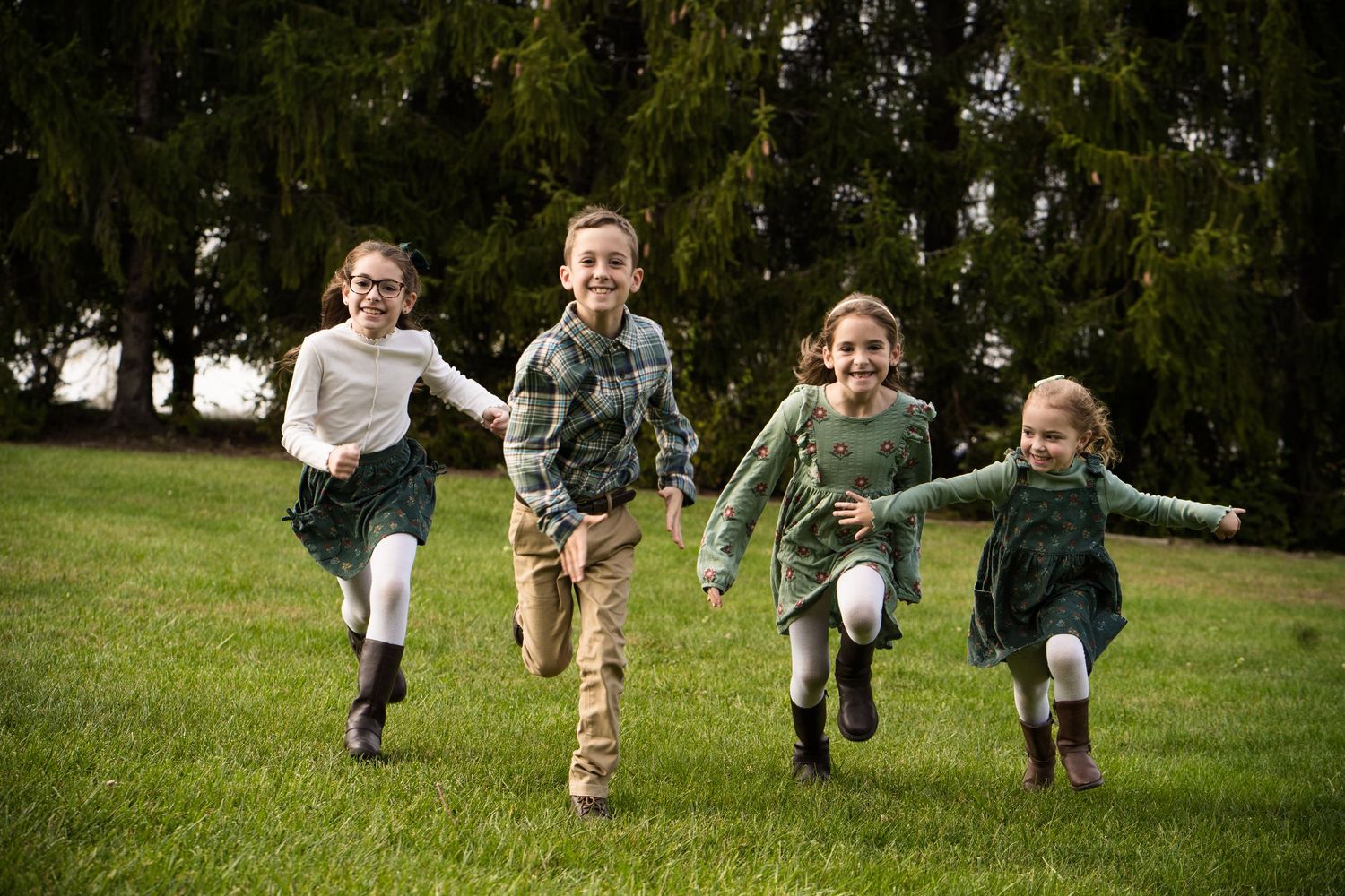 Four happy children running and laughing together on a green lawn with tall evergreen trees in the background.
