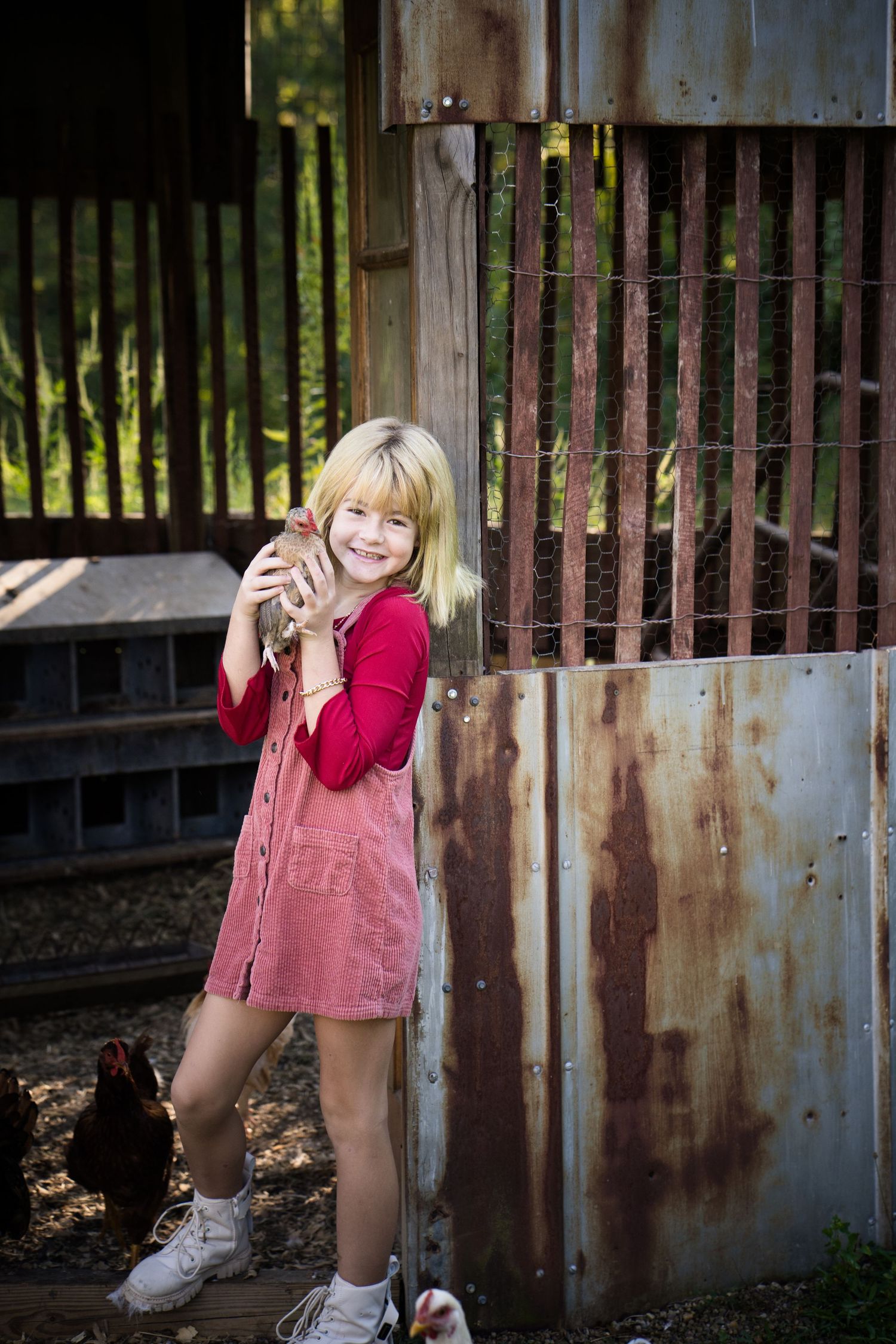 Smiling blonde girl in pink dress holding a chicken near a rustic barn with hens nearby.