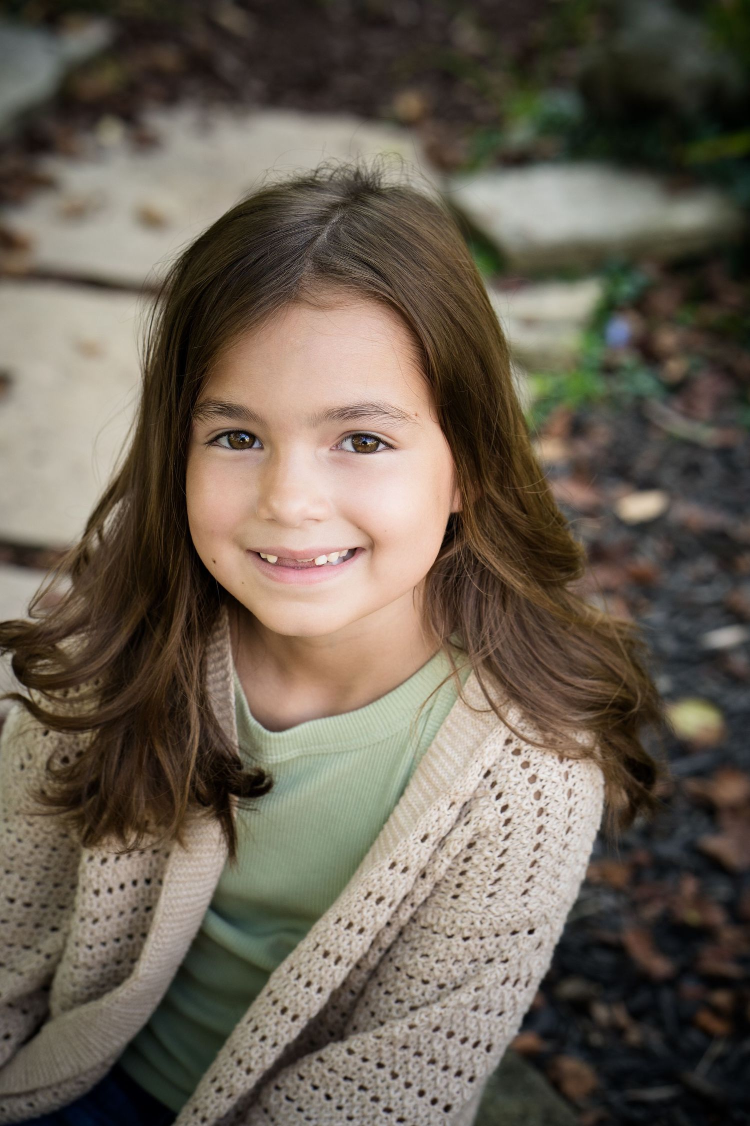 A smiling young girl with long brown hair wearing a beige knit cardigan and green shirt outdoors.