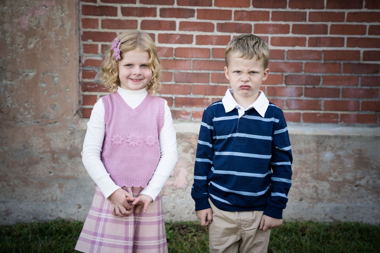 Two young children, a girl in pink dress and boy in striped shirt, posing together in front of a brick wall.