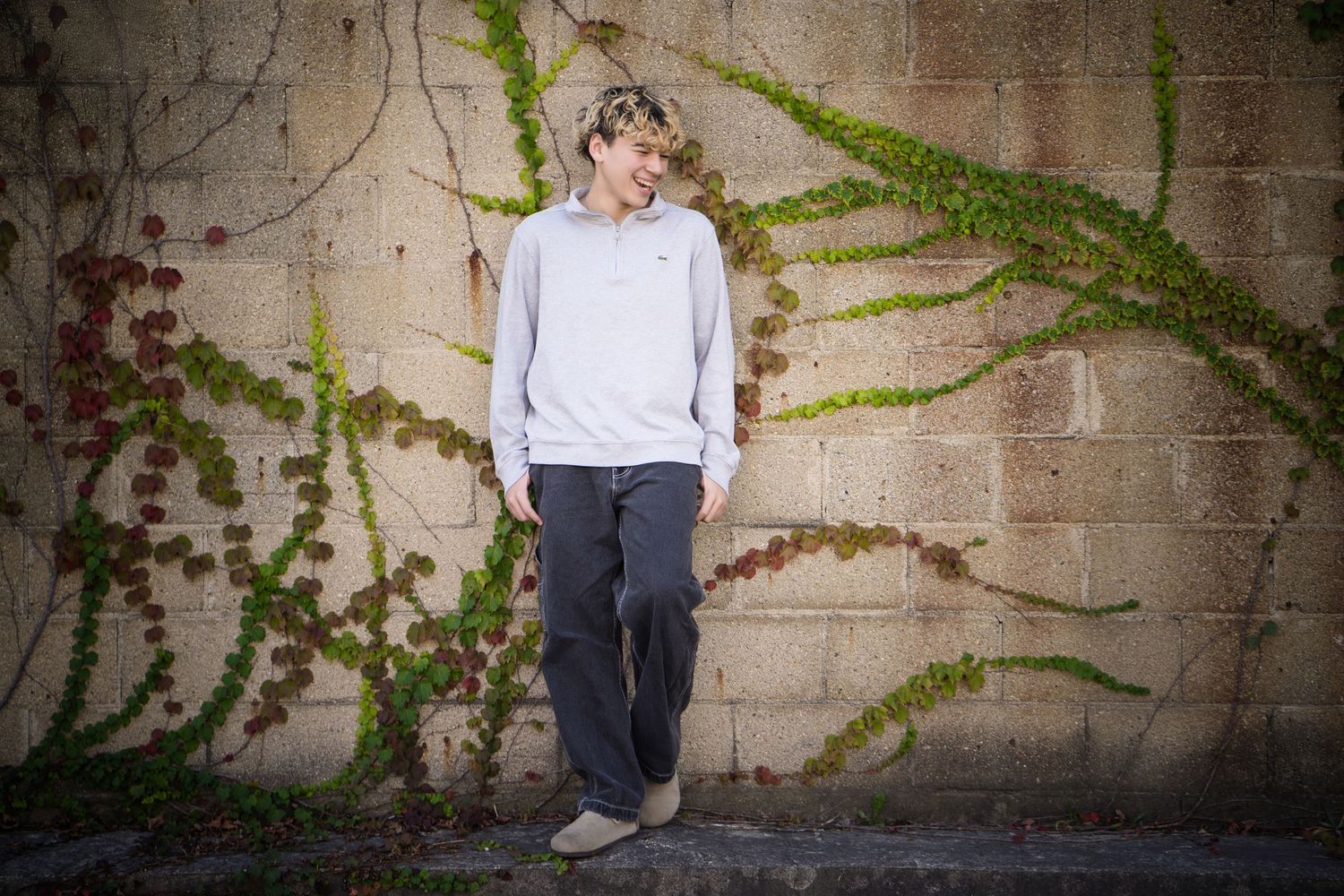Young man in gray sweatshirt and dark pants leaning against a stone wall with green ivy vines.
