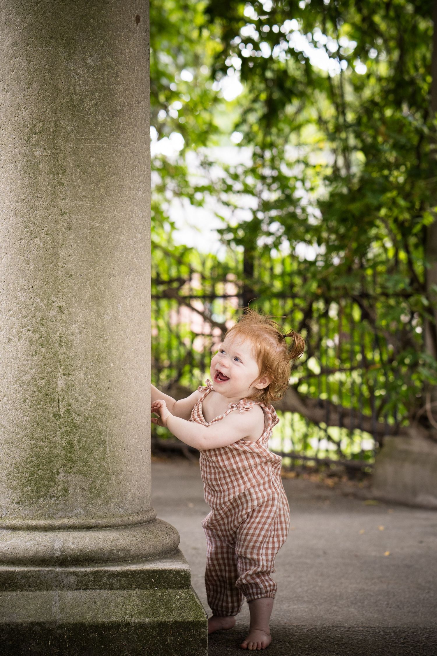 Happy red-haired toddler in plaid romper standing barefoot by stone column in lush green garden.