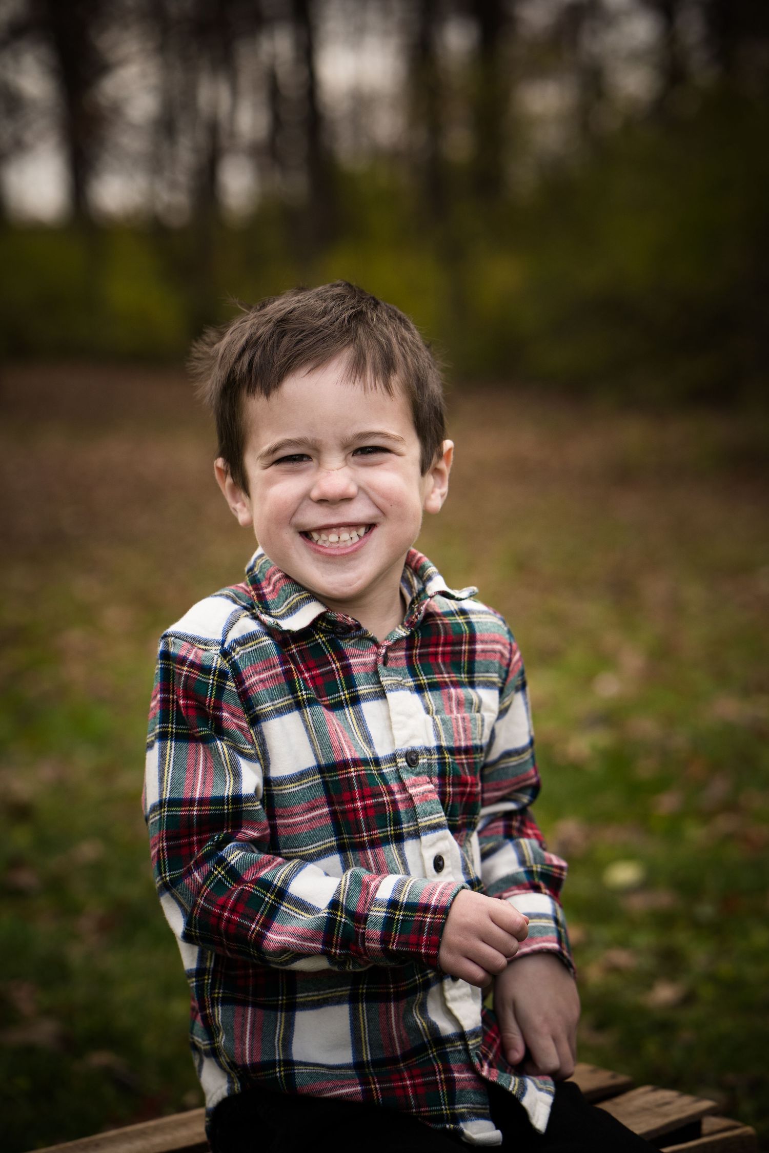 A smiling young boy in a plaid flannel shirt sits outdoors in a park setting with trees and greenery in the background.