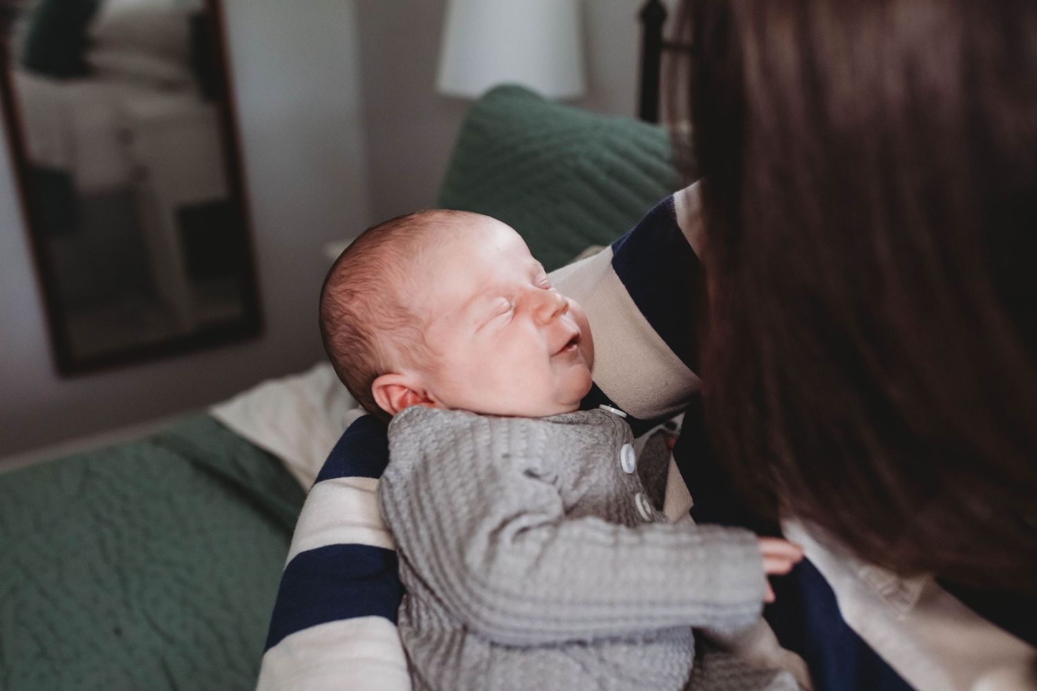 A mother gazes lovingly at her sleeping newborn baby dressed in a gray knit outfit.
