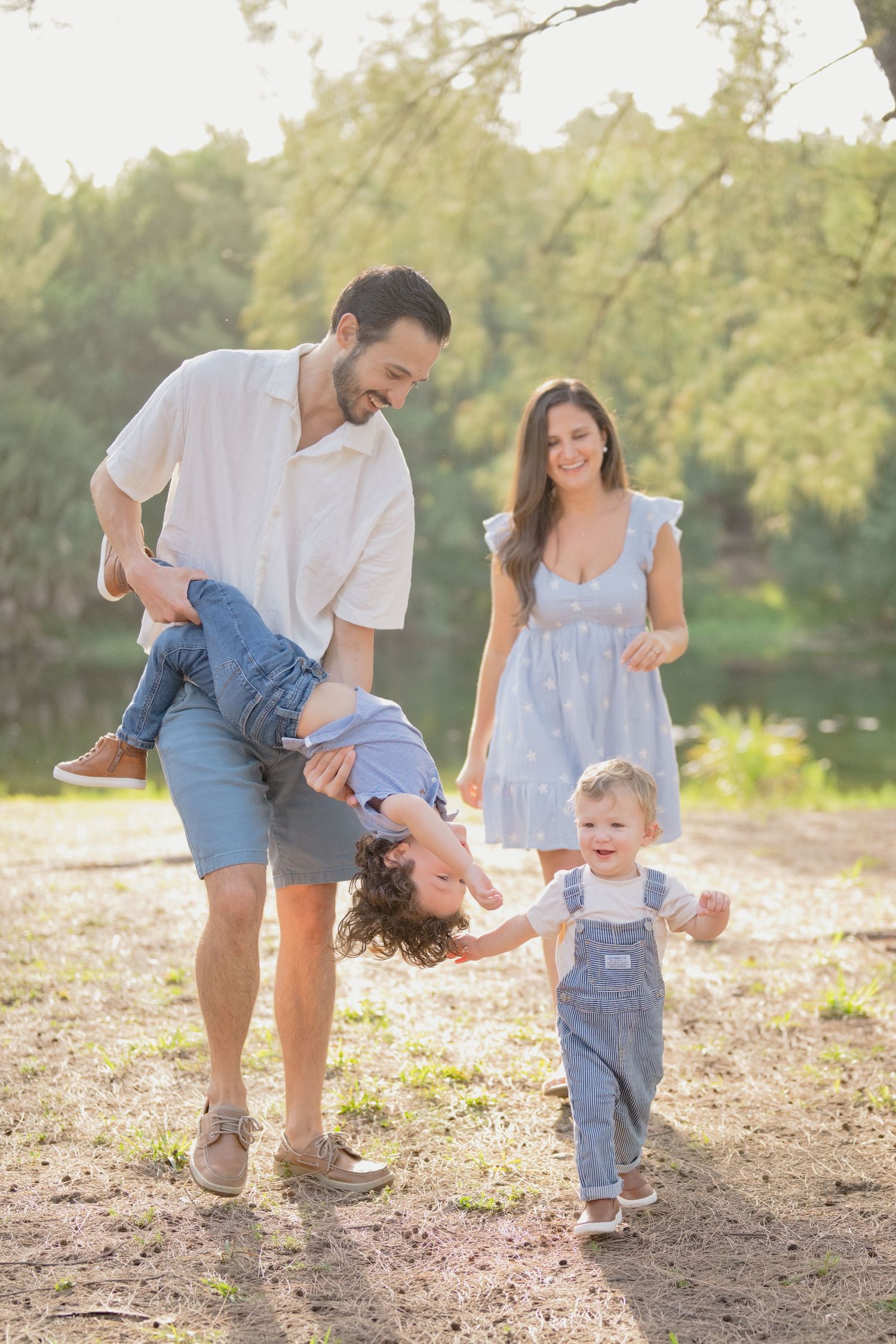 Family enjoying sunny day outdoors, dad swinging child upside down while mom watches toddler in overalls laugh.