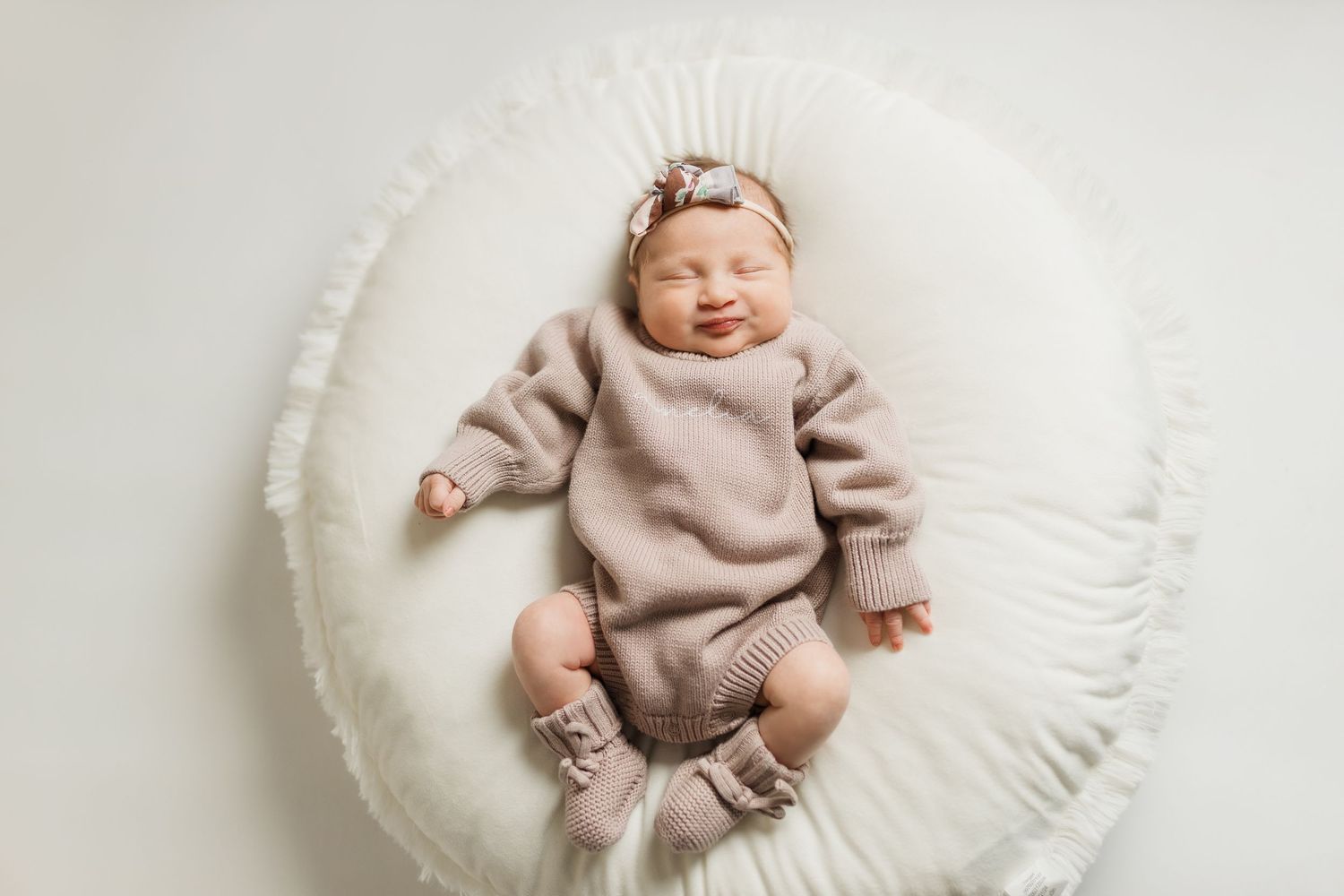 Newborn baby wearing soft pink knit outfit sleeps peacefully on round white cushion.