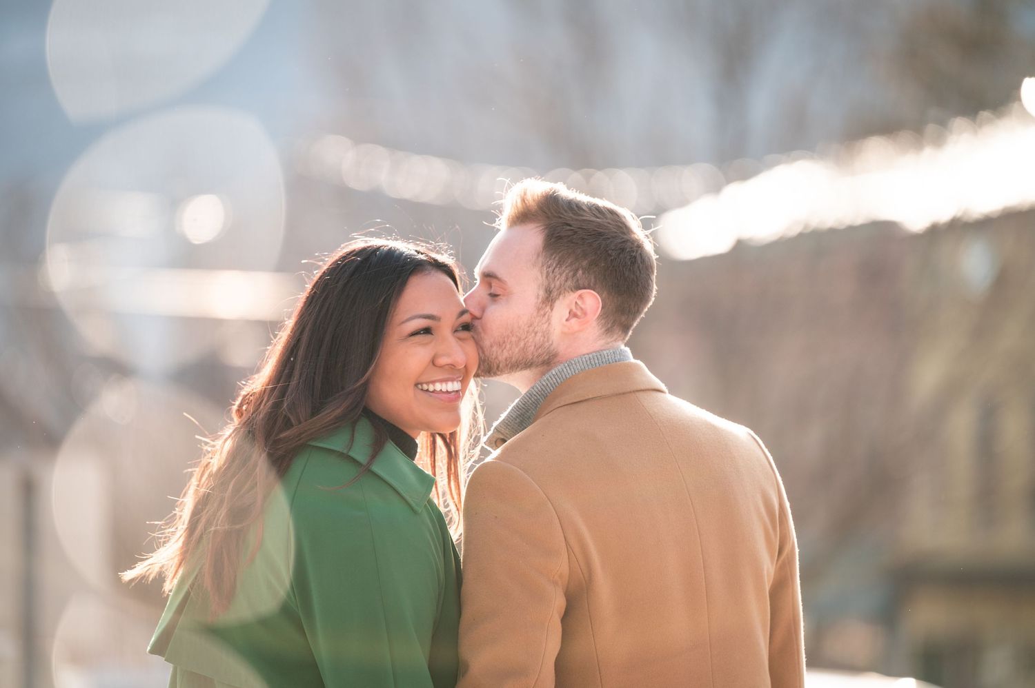 A couple in cozy winter coats share an intimate moment outdoors during golden hour, backlit by soft sunlight.