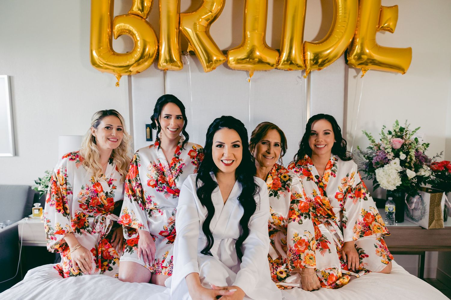 Group photo with gold BRIDE balloon letters and floral robes during wedding preparation.