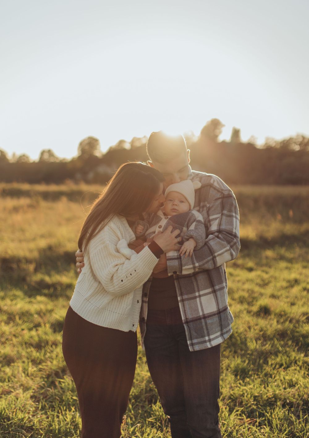 A couple cuddles with their baby in a sunlit field at golden hour.