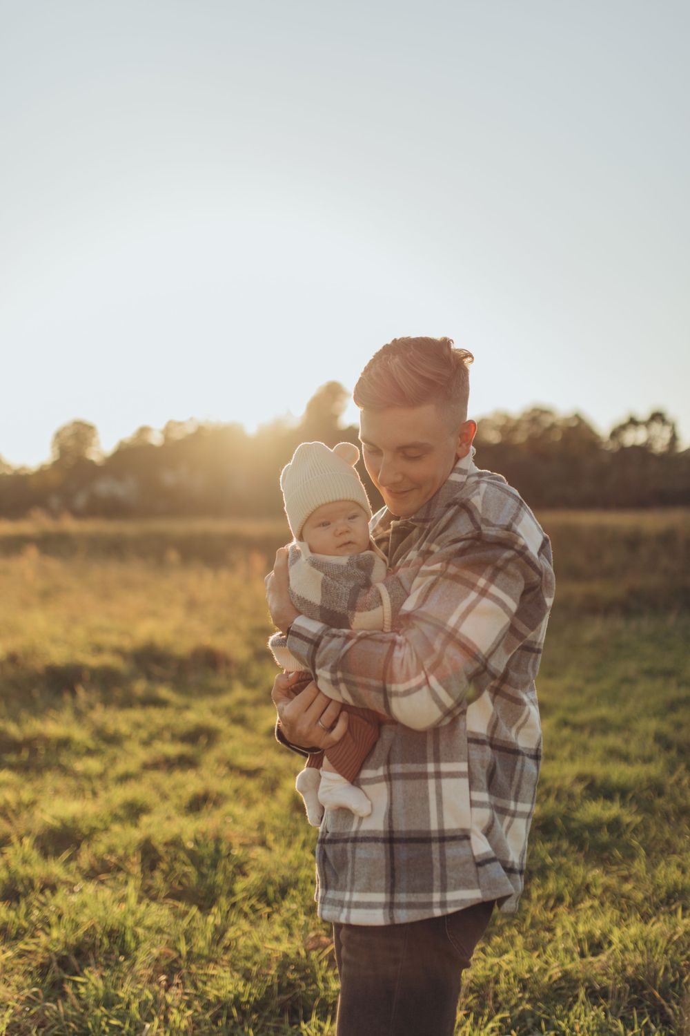 Person in plaid shirt holds baby against golden sunset in grassy field.