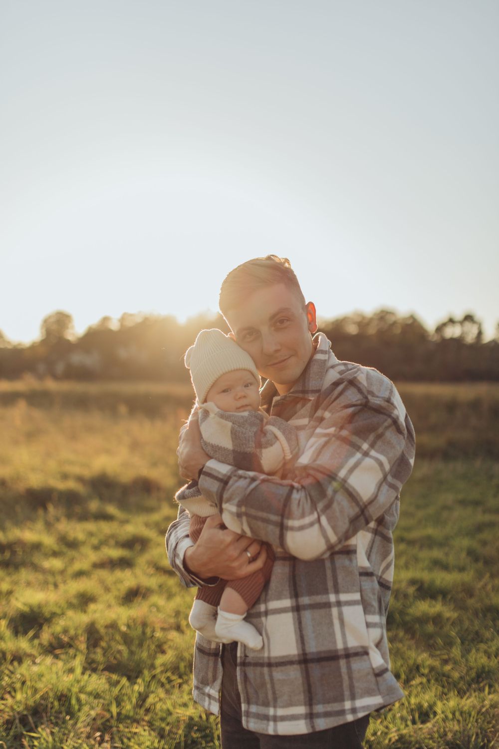 Person in plaid flannel cradles infant in sun-drenched field.