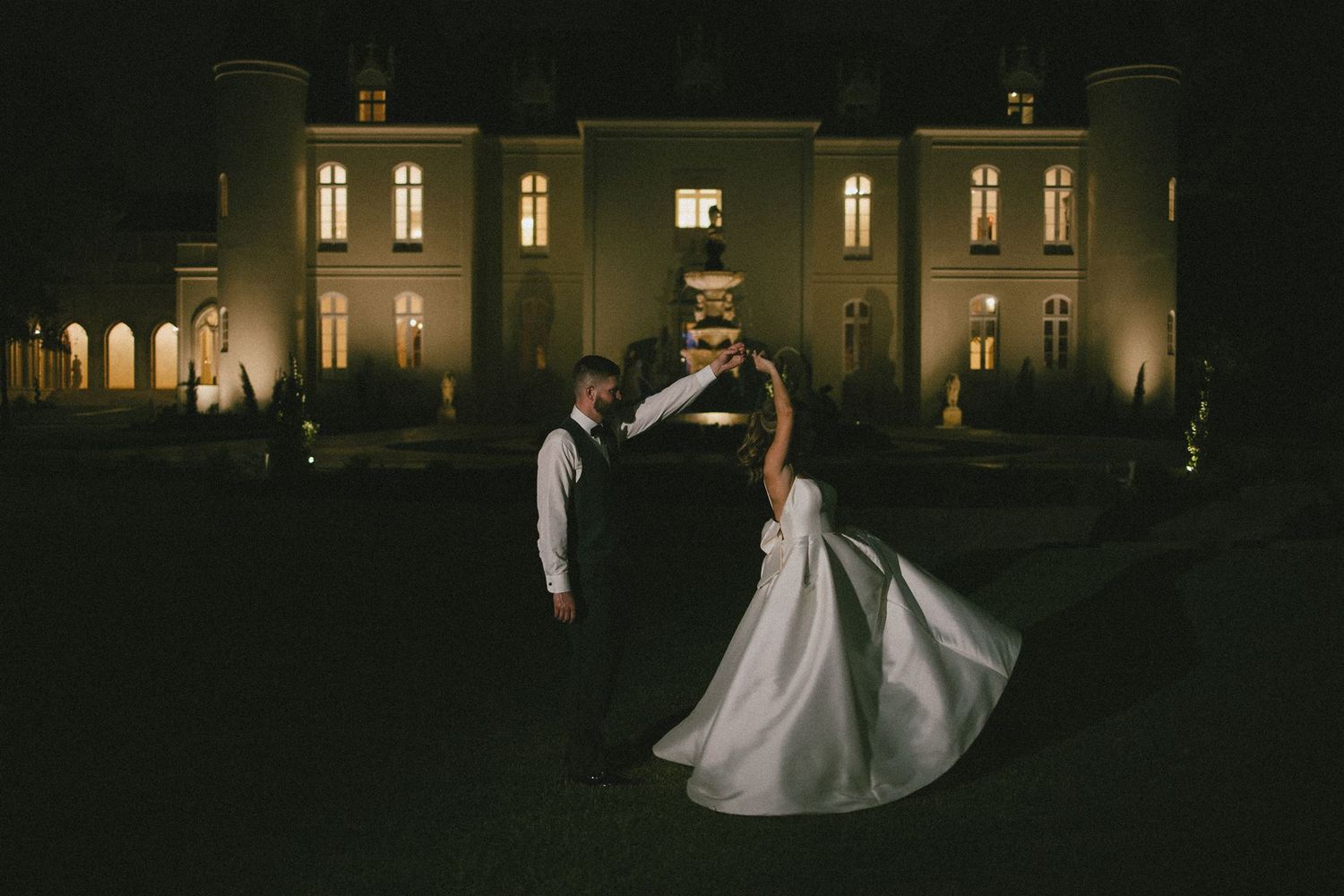 A romantic nighttime dip pose in front of an elegantly lit mansion with glowing windows and arched walkways.