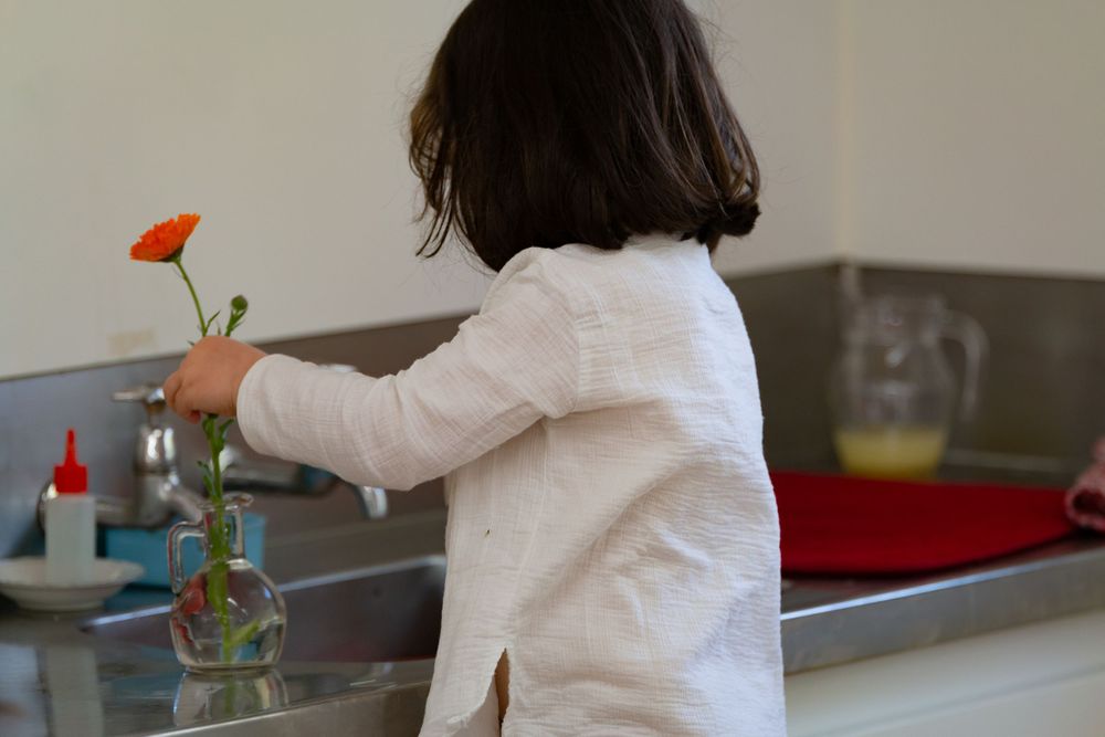 A person in a white top arranges a red flower in a glass vase at a kitchen counter.