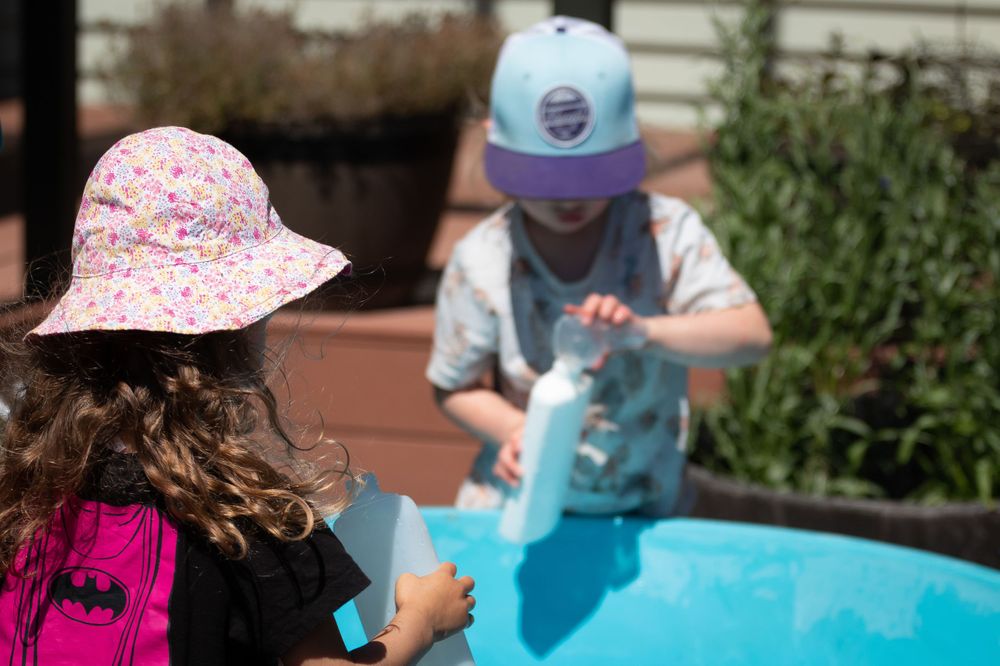 Two kids playing with blue water outdoors while wearing sun protection.