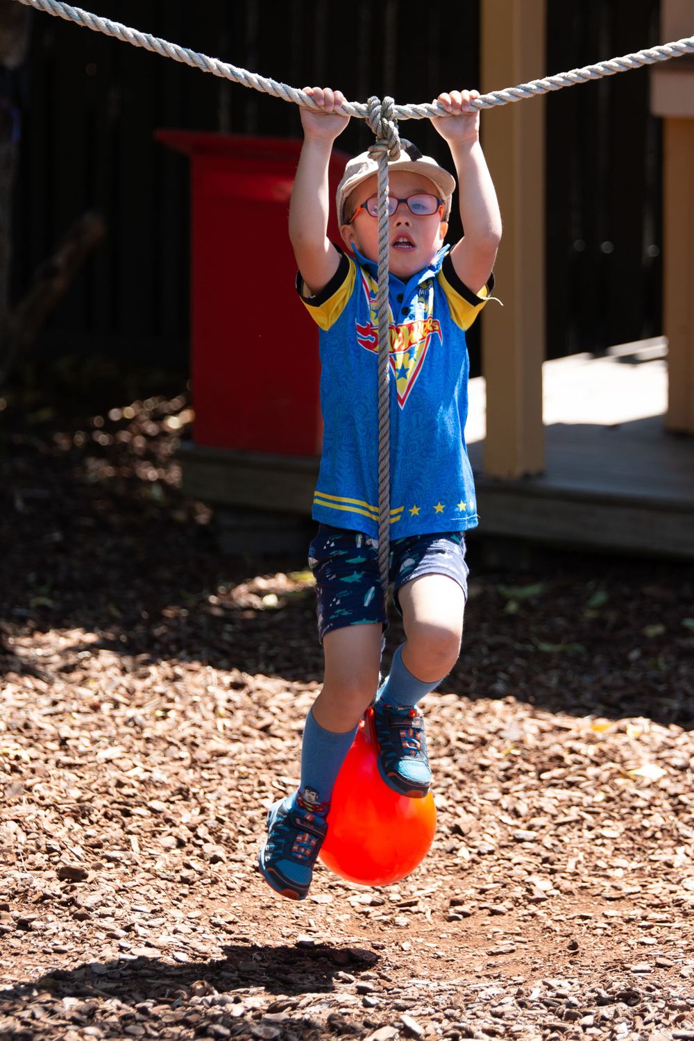 Child in superhero shirt and glasses traversing a rope obstacle course outdoors.