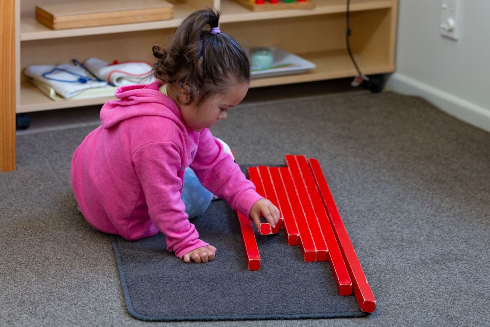 Young child in pink hoodie arranging red wooden blocks on gray carpet.