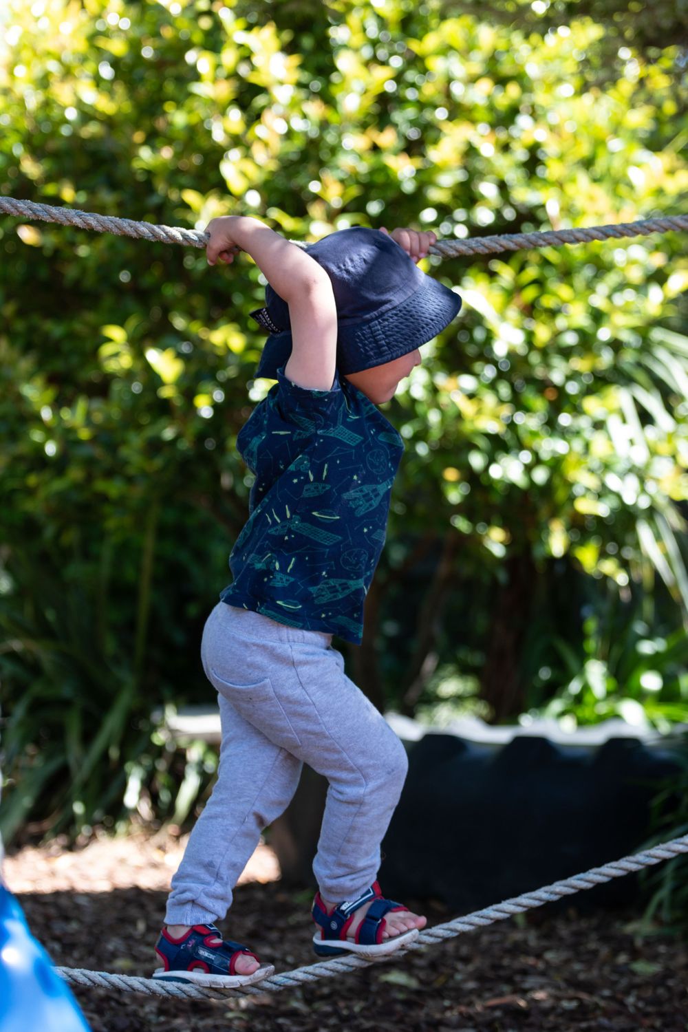 Child in navy outfit and hat balancing on a rope bridge in a garden setting.