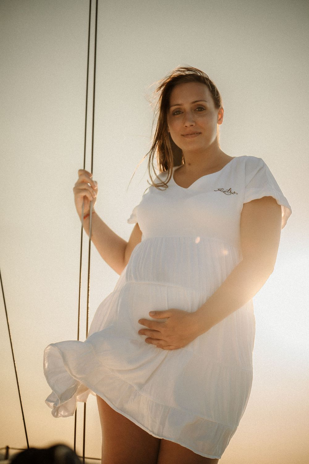 Woman in flowing white dress stands against sailing mast during sunset.