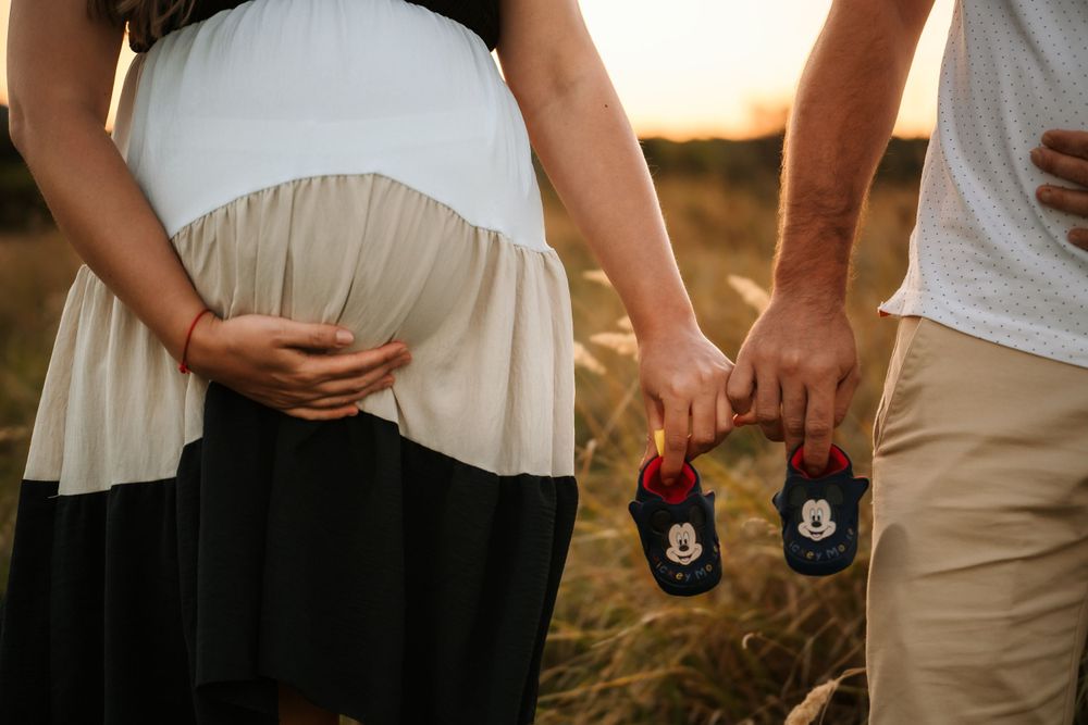 Expecting couple holds tiny baby shoes while standing in a field at sunset.