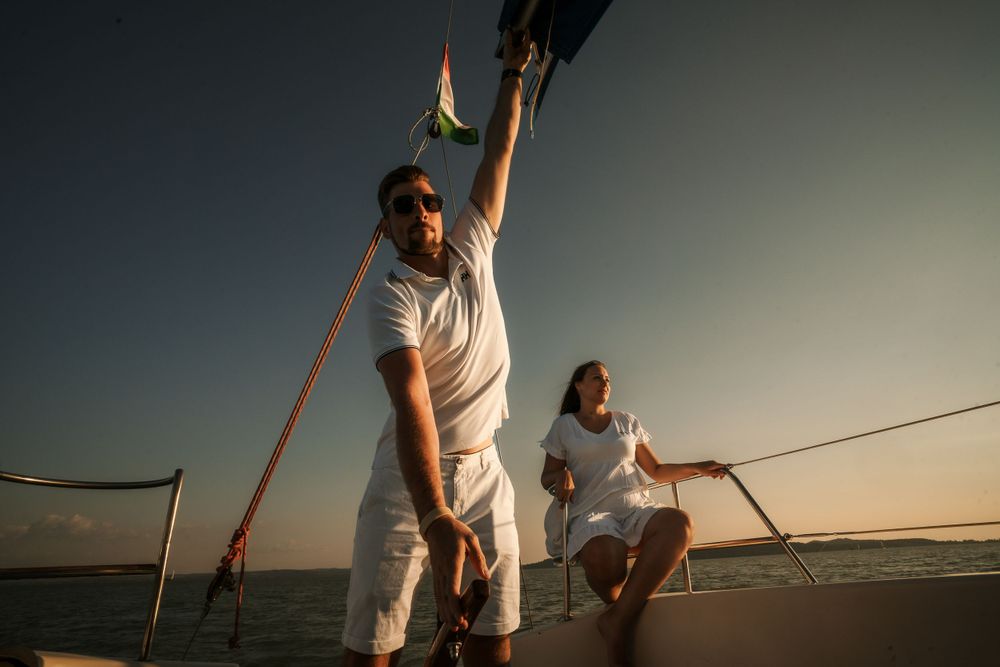 Two sailors in white attire managing ropes on a sailboat during golden hour.